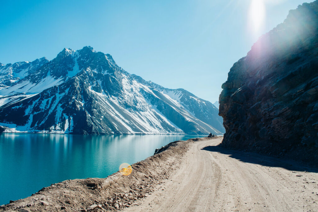 A winding dirt road curves along a bright turquoise lake beneath towering snow-capped Andes mountains, with sunlight hitting the rocky cliffside—capturing the dramatic landscapes near Embalse El Yeso, a stunning destination often included in what to do in Santiago for nature lovers.