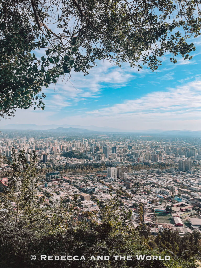 A wide panoramic view of Santiago’s dense skyline seen from an elevated lookout framed by leafy tree branches, with the Andes mountains faintly visible in the distance—one of the most popular things to do in Santiago from viewpoints like Cerro San Cristóbal, a Santiago top attraction.