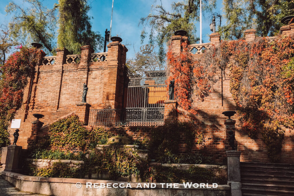 A historic brick wall covered in red and green ivy surrounds an ornate iron gate and small statues, with trees rising behind it under a blue sky—this is Cerro Santa Lucia, often included in lists of things to do in Santiago and Santiago top attractions.