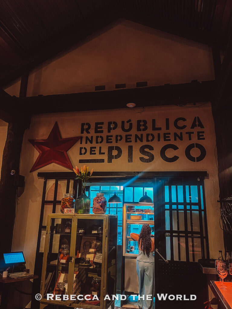A warmly lit bar interior with a sign reading “República Independiente del Pisco,” jars and bottles on display, and a person standing at a small kitchen window—highlighting unique nightlife experiences among Santiago top attractions.