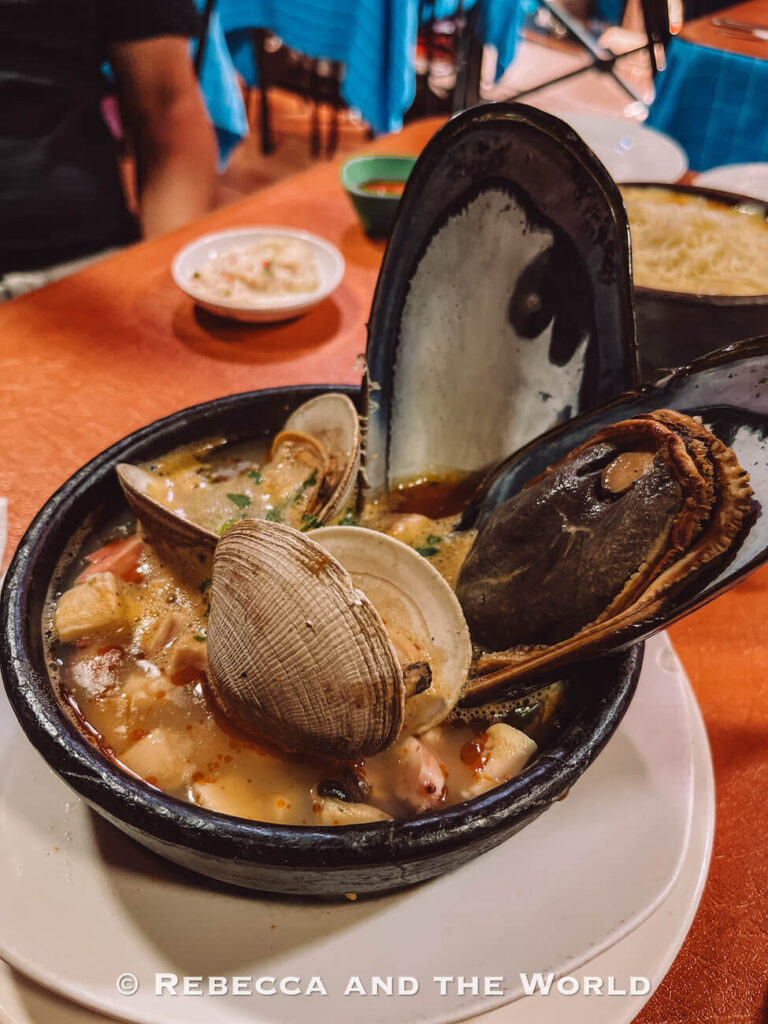 A steaming bowl of Chilean seafood stew filled with clams, mussels, and broth sits on a restaurant table—representing traditional cuisine and what to do in Santiago for food lovers, especially dishes commonly found at Mercado Central.