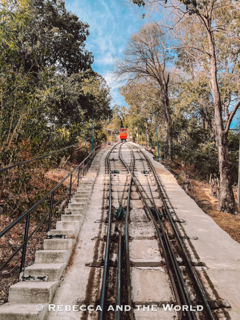 A red funicular car travels up steep tracks flanked by trees and a stairway, heading toward the summit of Cerro San Cristóbal—one of the most iconic things to do in Santiago for panoramic views and a classic Santiago top attraction.
