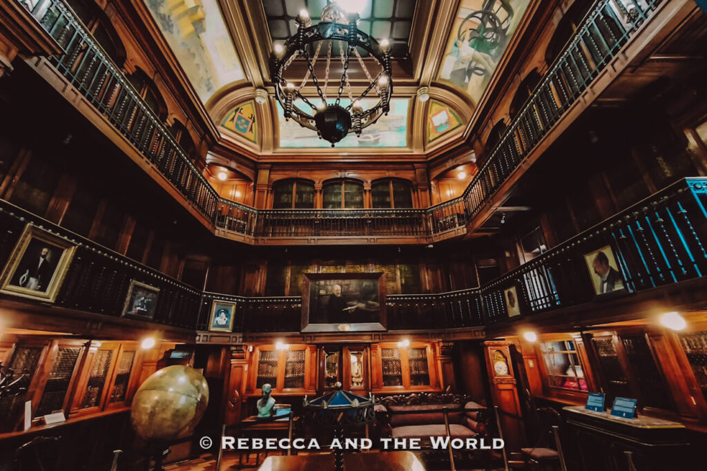 An ornate historic library interior with dark wooden shelves, a second-floor balcony, and a large chandelier hanging from a stained-glass ceiling, with portraits, antique furniture, and a globe on display—likely the Biblioteca Nacional de Chile in Santiago, showcasing cultural heritage and one of the more unique Santiago top attractions for those exploring what to do in Santiago.