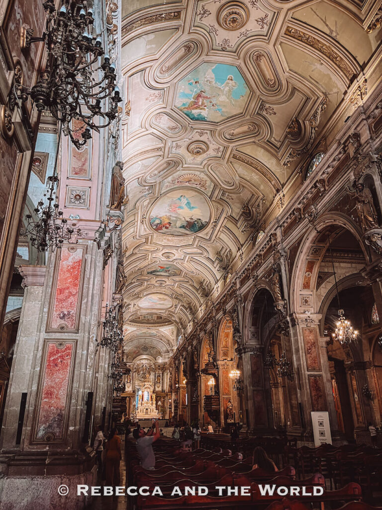 An ornate cathedral interior with high vaulted ceilings, detailed frescoes, chandeliers, and rows of wooden pews, likely inside the Metropolitan Cathedral of Santiago—highlighting religious architecture among the top attractions in Santiago.