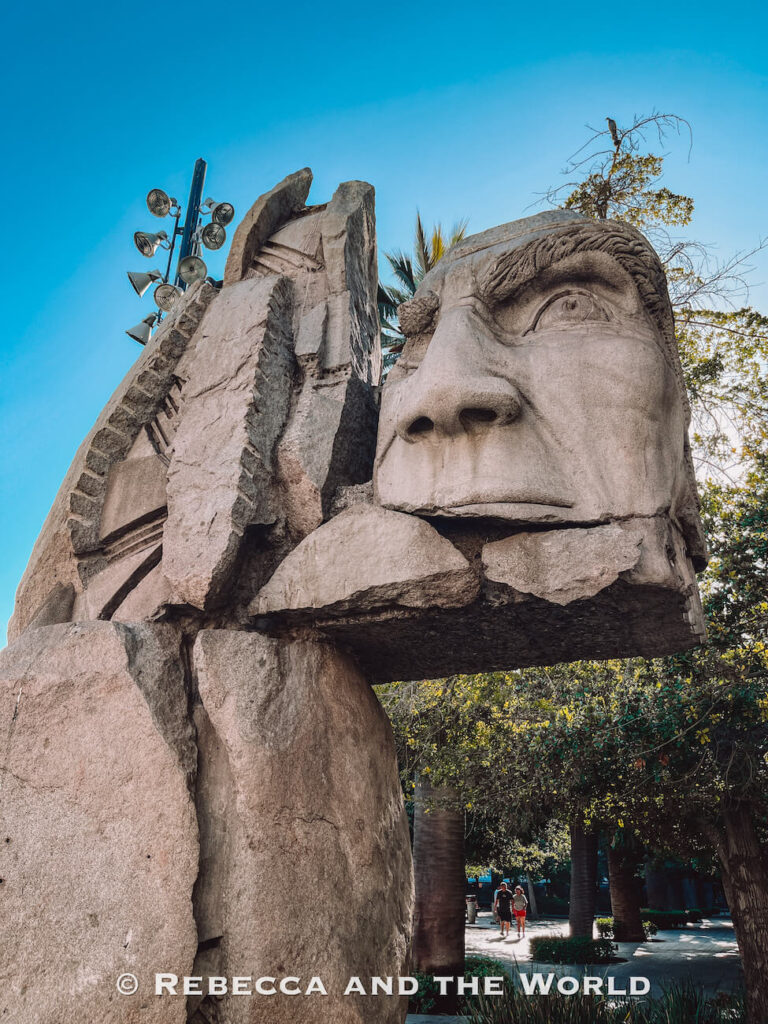 A large abstract stone sculpture featuring a carved human face stands in a leafy park setting, with people walking in the background—an example of public art found in places like Plaza de Armas, adding variety to things to do in Santiago. This is the Monument to Indigenous Peoples.