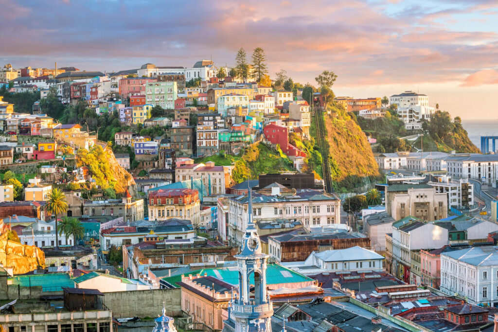 Colorful houses stacked along steep hillsides in Valparaíso glow in warm sunset light, with narrow streets, historic buildings, and a funicular track visible—highlighting a popular coastal day trip and one of the most scenic things to do in Santiago beyond the city.