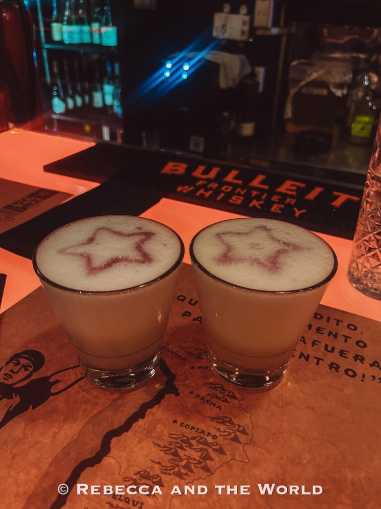 Two pisco sour cocktails with star-shaped foam designs sit on a bar counter with bottles and dim lighting in the background, illustrating what to do in Santiago for nightlife and local drinks.