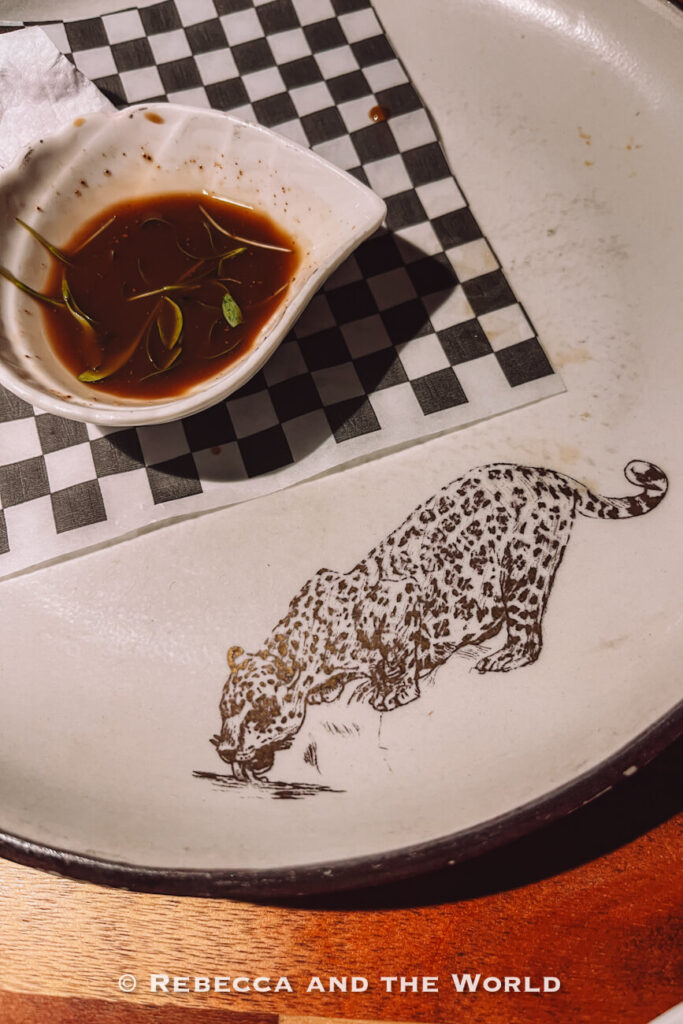 Small dish of dark dipping sauce with herb leaves beside a large ceramic plate decorated with a spotted wild cat illustration, photographed at a Bogotá restaurant featured in a 2 days in Bogotá itinerary.