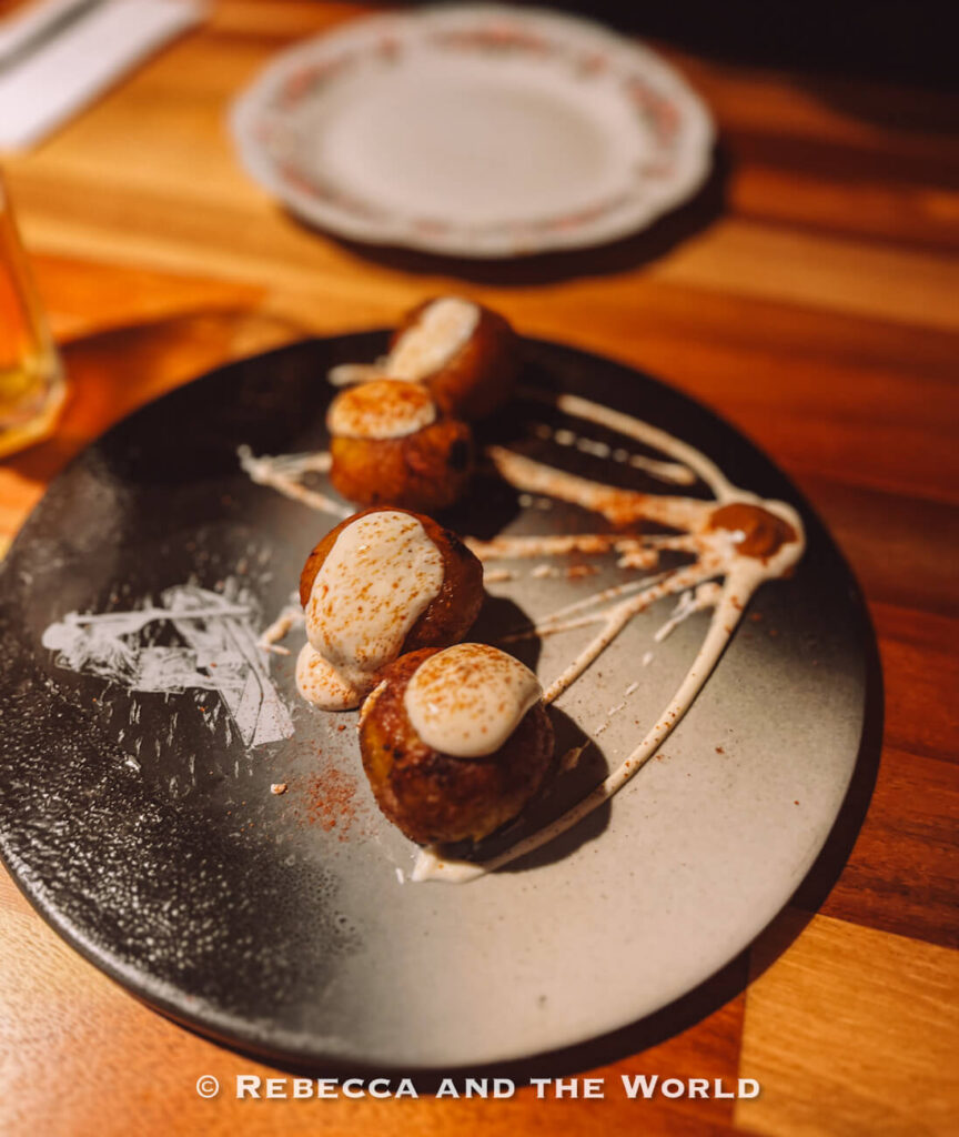 Four golden-brown croquettes topped with creamy white sauce and arranged on a black plate with artistic sauce streaks, showcasing local food as one of the things to do in Bogotá.