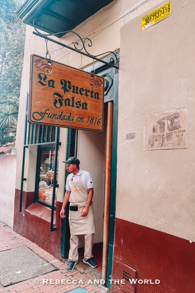 Restaurant entrance with a wooden sign reading “La Puerta Falsa, Fundada en 1816” and an employee standing in the doorway of the historic building in Bogotá’s old town, featured in a Bogotá travel guide.