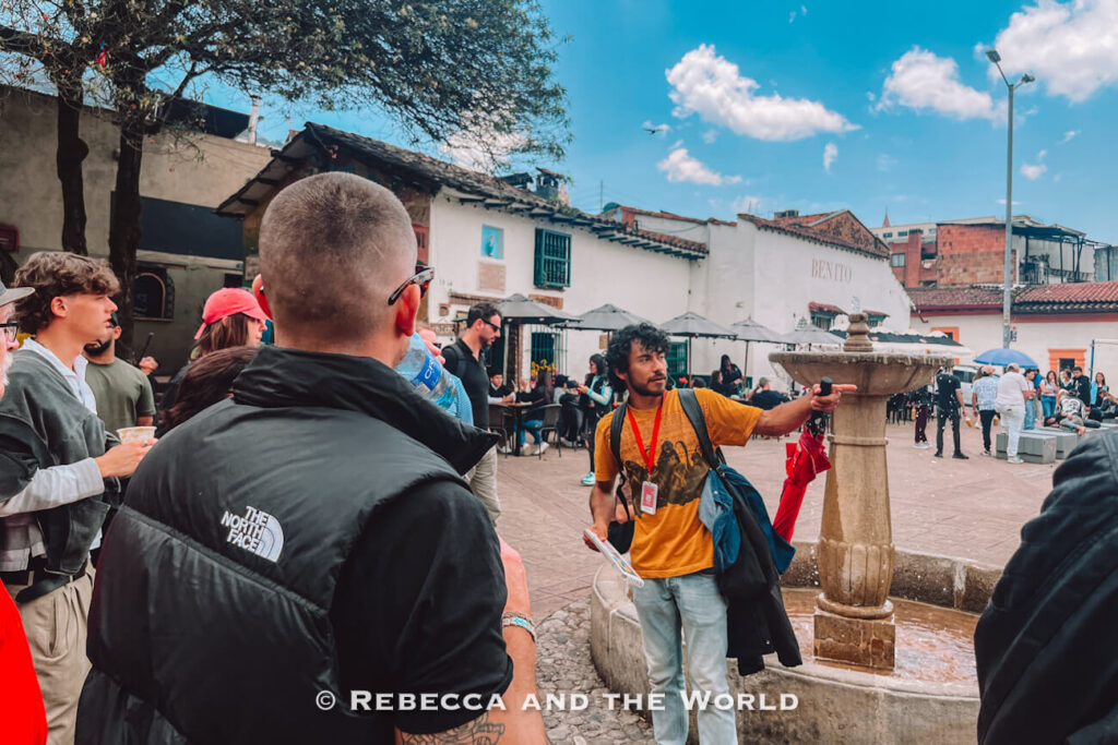Tour guide speaking beside a stone fountain while a group of visitors gathers around in a busy Bogotá plaza, one of the popular things to do in Bogotá.