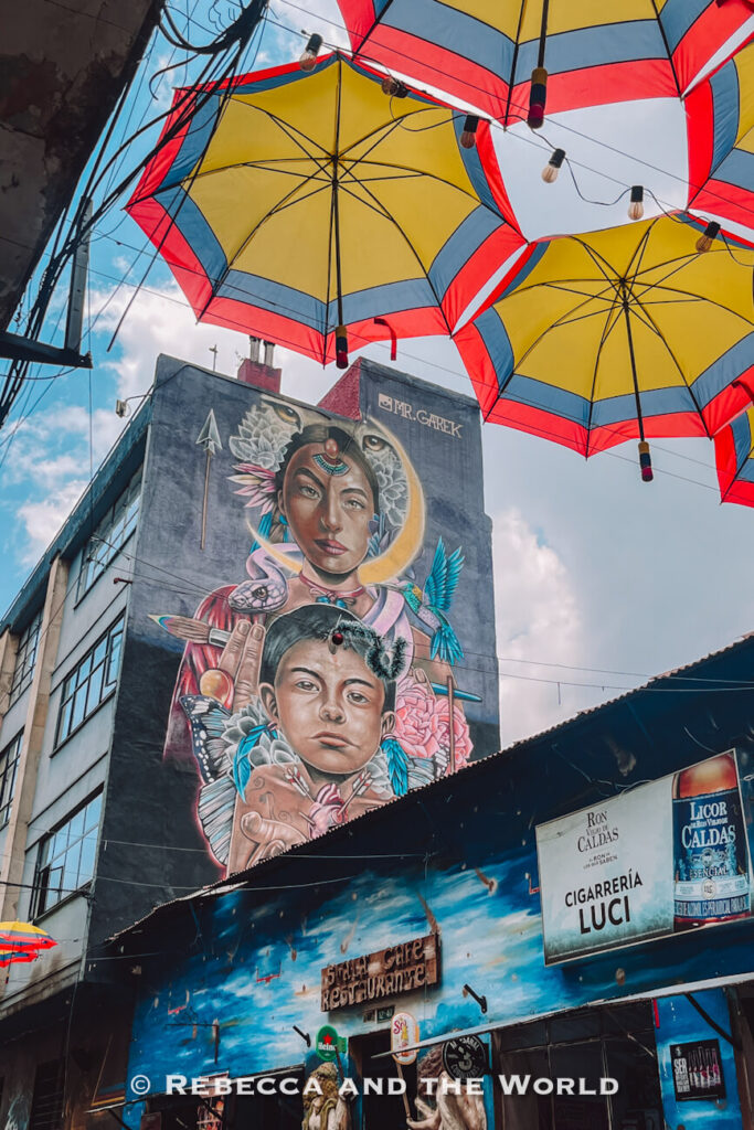 Large mural of two figures surrounded by flowers, birds, and symbolic artwork on the side of a city building beneath colorful umbrellas, featured in a Bogotá travel guide.