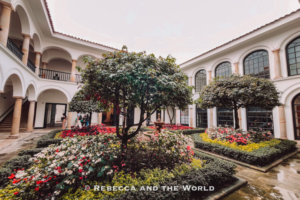 Formal courtyard garden with trimmed hedges, flowering plants, and a central fountain surrounded by a white colonial building - the Museo Botero- a peaceful stop on a Bogotá itinerary.
