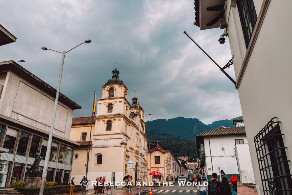 Historic church with twin towers set against forested mountains and dark storm clouds, viewed from a city street in Bogotá, featured in a Bogotá travel guide.