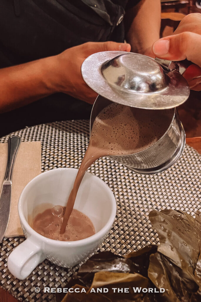 Person pouring a frothy hot chocolate from a metal pitcher into a white cup at a restaurant table, showcasing traditional Colombian food in a Bogotá travel guide.