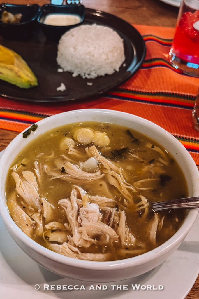 Bowl of ajiaco chicken soup with shredded chicken, potatoes, and corn served with rice and avocado on the side, highlighting local cuisine in a Bogotá itinerary.