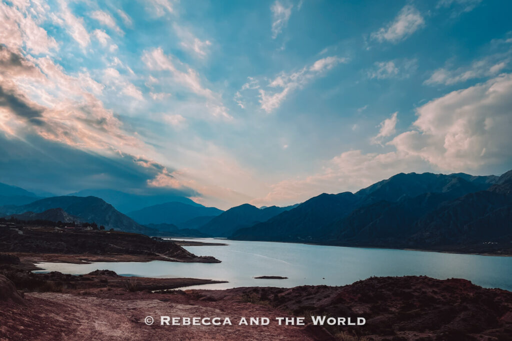 Wide view of a calm lake surrounded by dark Andes mountains, with rocky shoreline in the foreground and dramatic blue sky filled with soft clouds and warm sunset light; a scenic landscape for a 3 days in Mendoza itinerary.