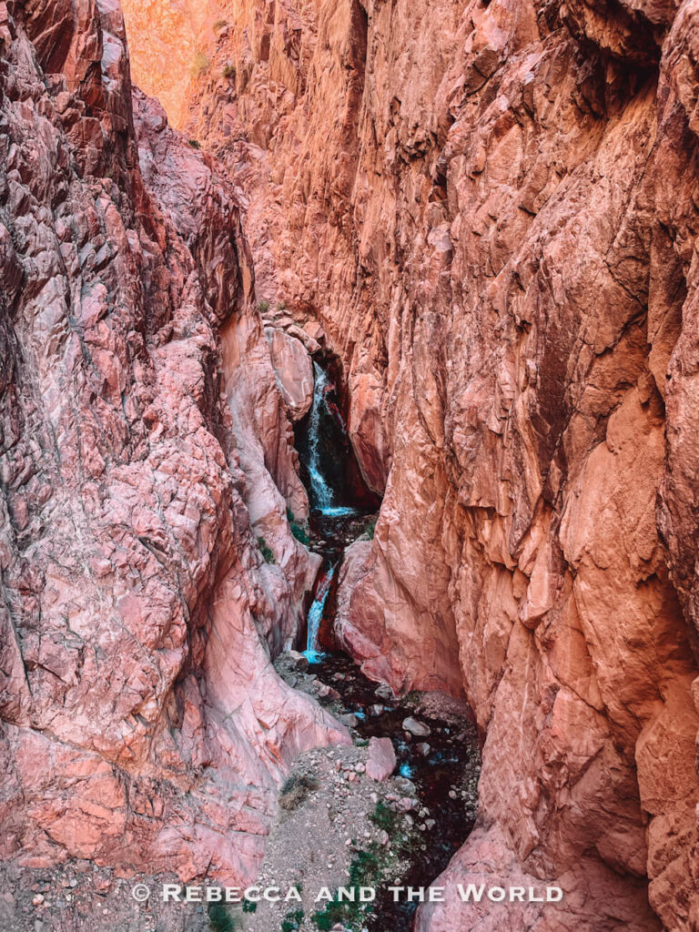 View looking down into a narrow reddish rock gorge with a small stream and waterfall flowing through the canyon floor. A dramatic stop for a Mendoza itinerary.