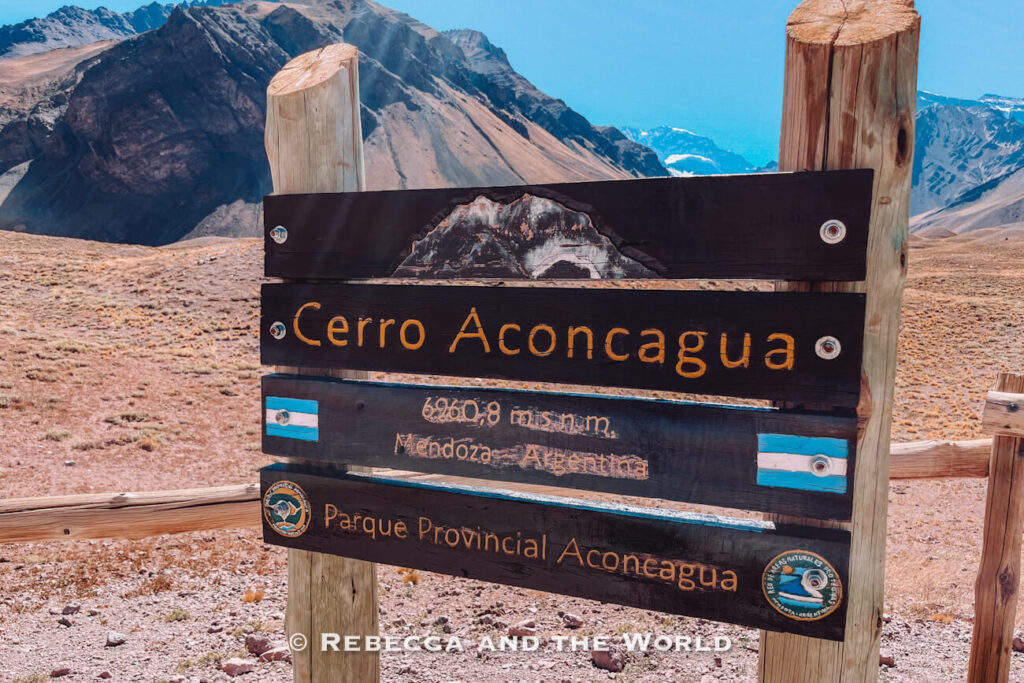 Wooden sign reading Cerro Aconcagua with mountain peaks visible in the background under bright sunlight. A classic answer to what to do in Mendoza.