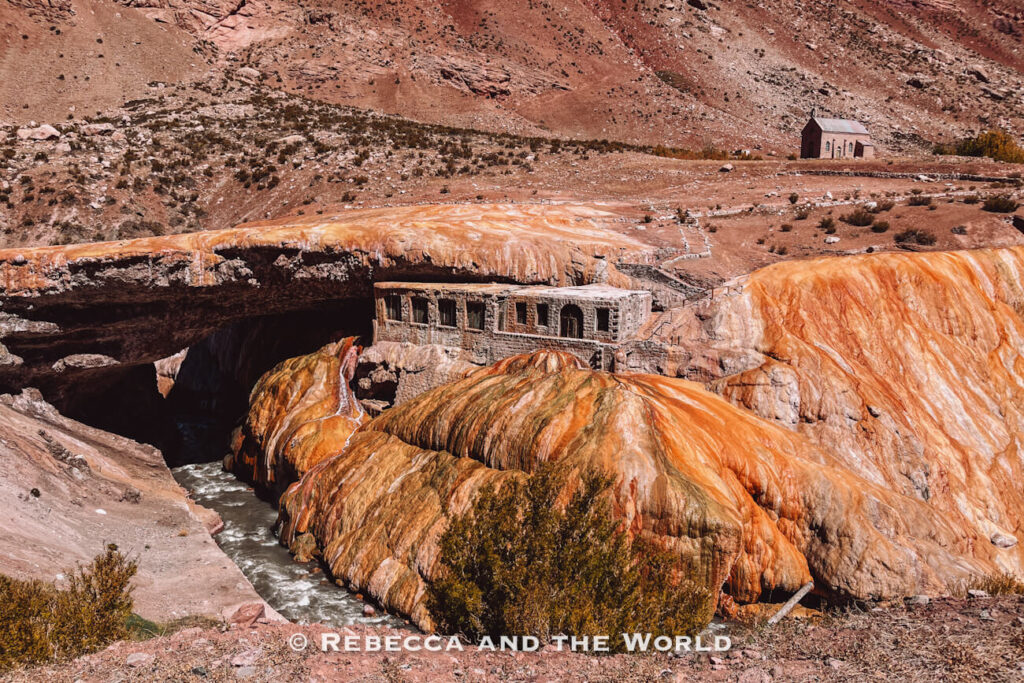 Mineral-covered orange rock formation with a stone building built into the hillside and water flowing underneath. The Puente del Inca is a unique stop among things to do in Mendoza.