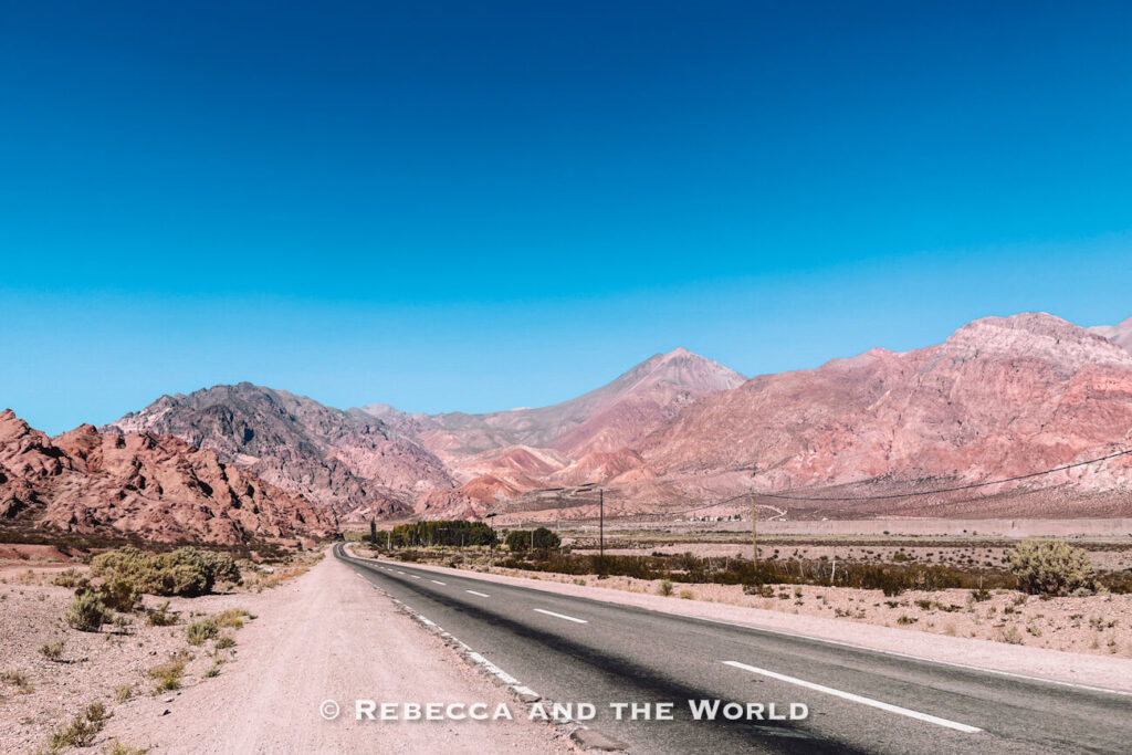 Empty paved road stretching through a wide valley of colorful rocky mountains under a clear blue sky. A scenic route for a Mendoza itinerary.