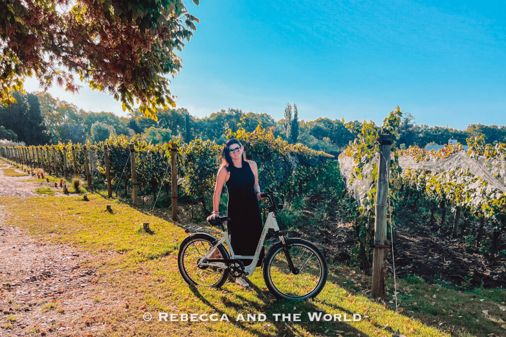A woman (the author of this article) standing with a bicycle between vineyard rows on a sunny day with trees and green grass surrounding the path. Wondering what to do in Mendoza? Grab a bike a cycle to different vineyards.