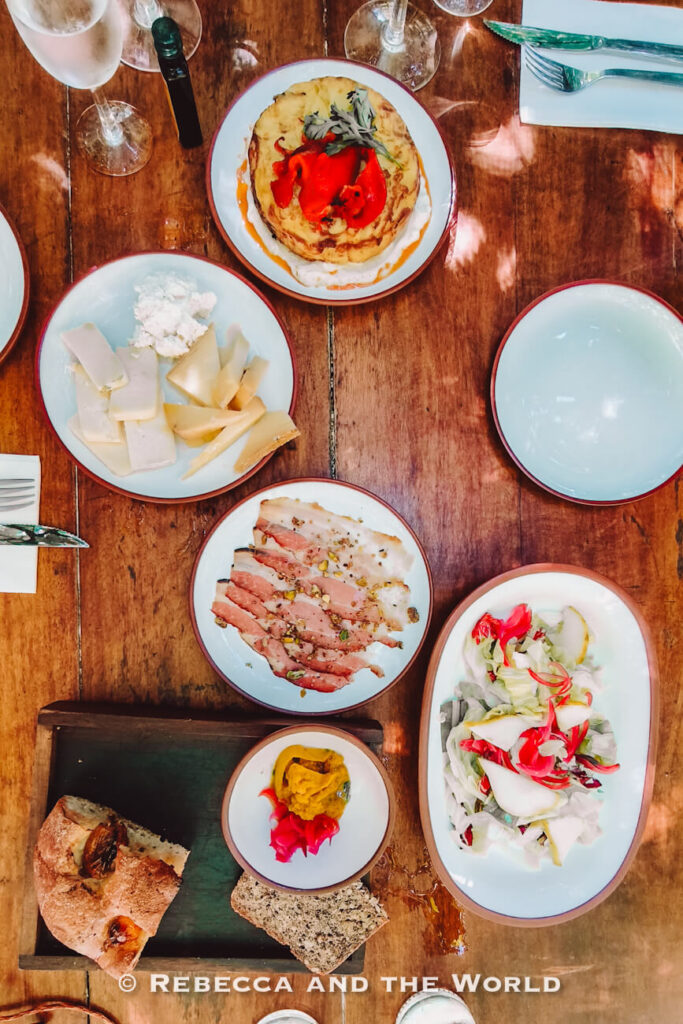 Overhead view of a wooden table set with cheeses, sliced meat, salad, bread, and plated dishes outdoors.