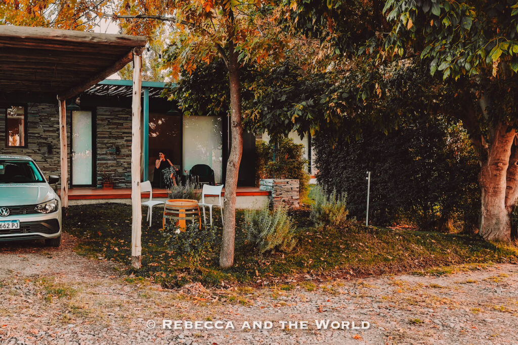 Small modern cabin surrounded by mature trees and autumn leaves, with a gravel driveway, parked white car, shaded patio seating, and a person sitting in the doorway relaxing outdoors. A tranquil accommodation setting and inspiration for what to do in Mendoza.