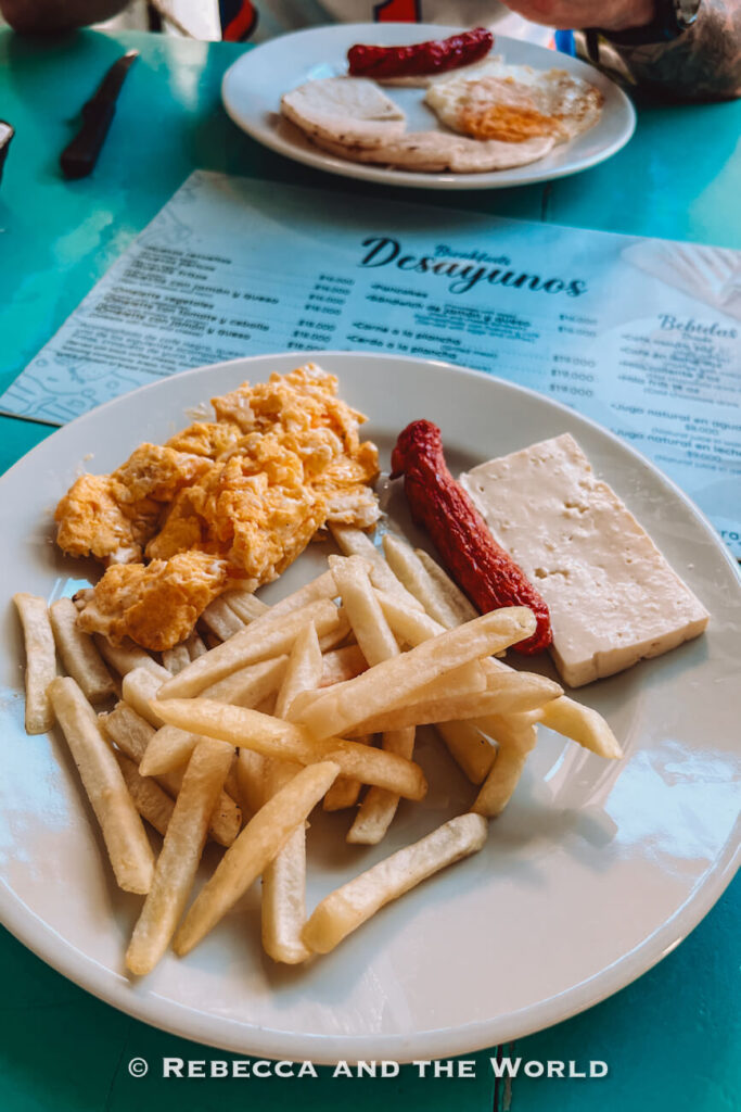 A plate of scrambled eggs, French fries, grilled sausage, and white cheese sits on a turquoise table, with another breakfast plate visible in the background. Sampling local breakfasts can be part of any Cartagena itinerary.