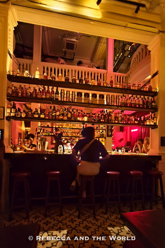 A dimly lit bar with tall shelves lined with liquor bottles, bartenders working behind the counter, and a person sitting on a stool facing the bar. An upper-level balcony overlooks the space, illuminated by warm amber and pink lighting. One of the most atmospheric things to do in Cartagena is exploring its cocktail bars.
