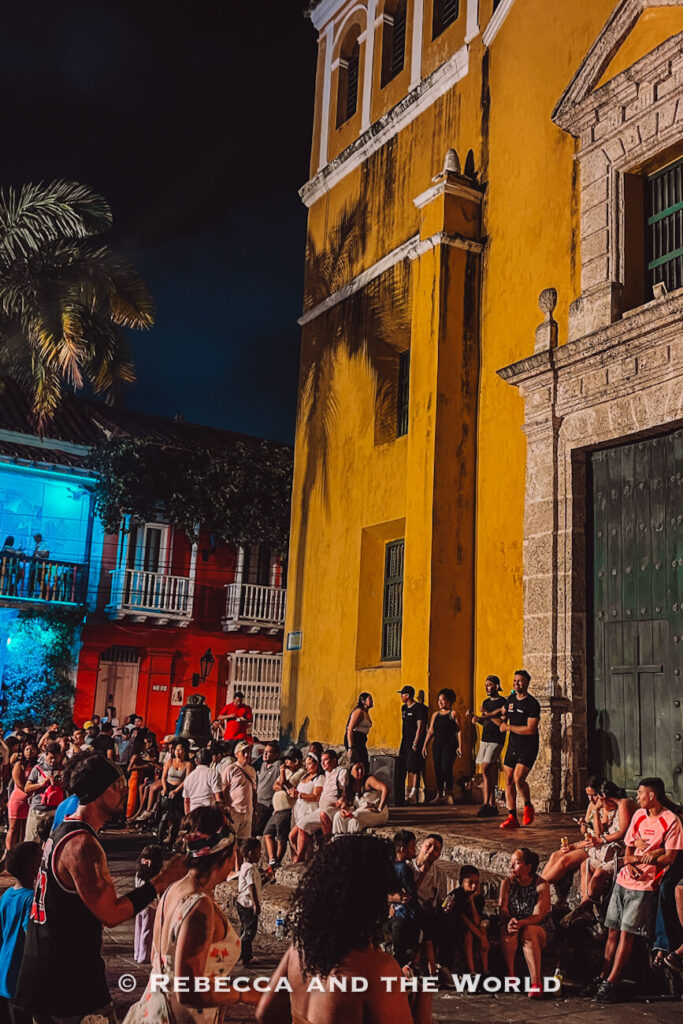 A large crowd gathers outside a yellow colonial church at night, with people sitting on steps and standing along the plaza. Watching local nightlife unfold is a great way to experience what to do in Cartagena.