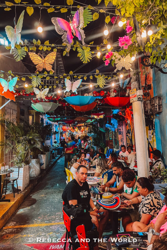A narrow alley filled with outdoor restaurant seating is decorated with hanging butterflies, umbrellas, string lights, and tropical plants. This lively atmosphere of Getsemani often appears in a Cartagena travel guide.