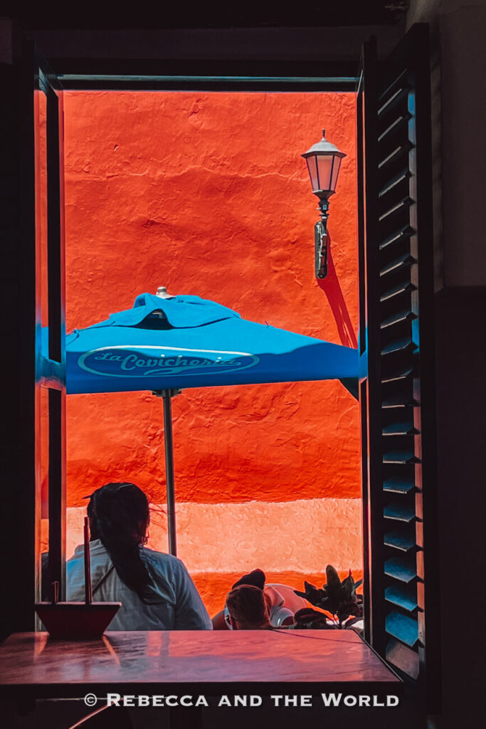 An open window frames a bright orange wall outside, a black street lamp, and a blue patio umbrella, while a seated person is partially visible below. The colorful architecture is one reason many visitors spend 3 days in Cartagena.