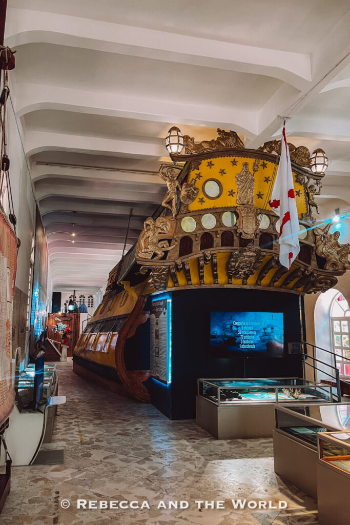 A large wooden ship replica fills the center of a museum gallery, surrounded by display cases and informational exhibits. Visiting the Museo Naval del Caribe is a worthwhile addition to a Cartagena itinerary.
