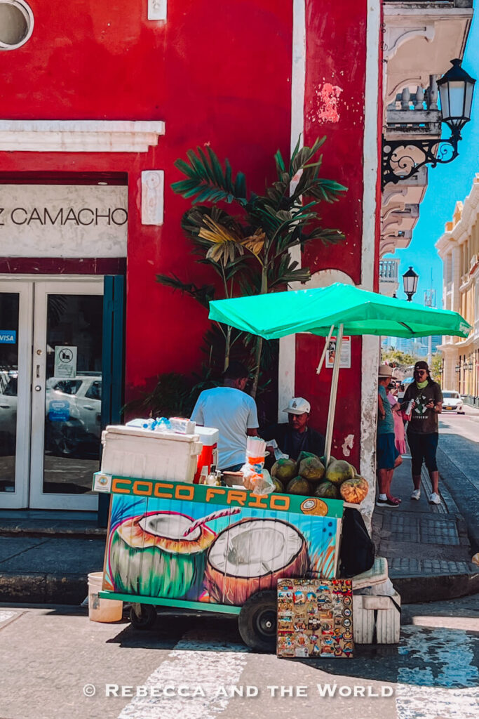 A street vendor stands beside a colorful cart selling fresh coconuts under a green umbrella at a street corner lined with colonial buildings. Sampling fresh coconut water is a popular answer to what to do in Cartagena.