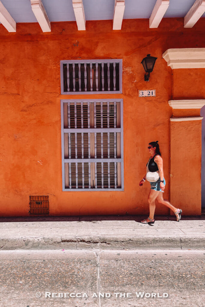 A person (the author of this article) walks past a bright orange colonial building with shuttered windows and a black wall-mounted lantern. The colorful streets are frequently featured in every Cartagena travel guide.