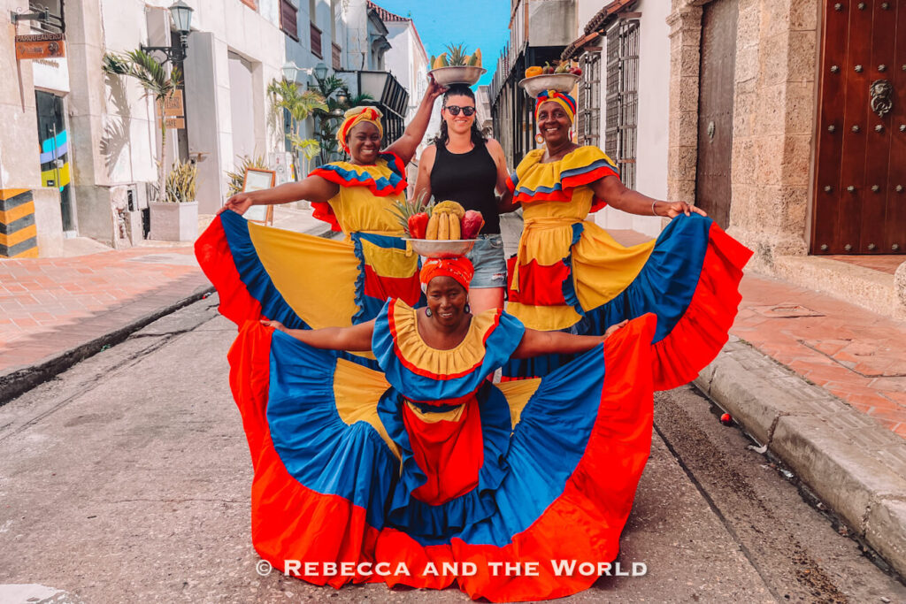 Three women - called palenqueras -wearing bright yellow, blue, and red traditional dresses pose around a woman (the author of this article) in the middle of a cobblestone street. Taking photos with local performers is a common activity during 3 days in Cartagena.