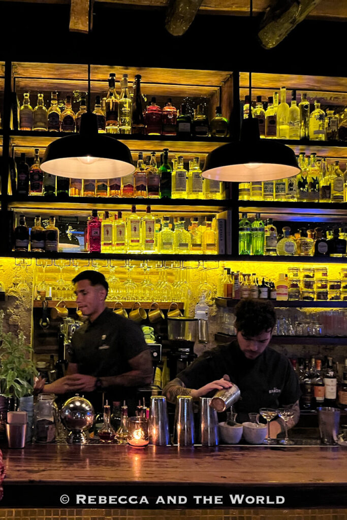 Two bartenders prepare drinks behind a bar backed by illuminated shelves filled with bottles and glassware. This is El Baron bar. Cartagena’s cocktail scene is often highlighted in a Cartagena itinerary.
