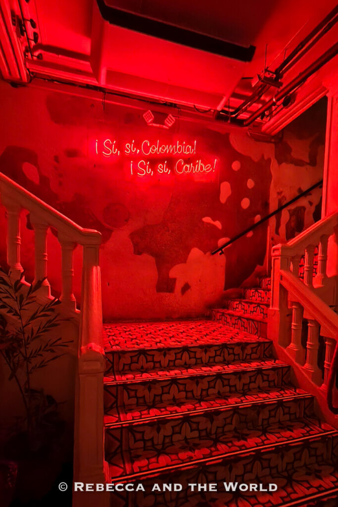 A staircase with patterned tile steps leads upward between white railings toward a red neon sign reading “¡Sí, sí, Colombia! ¡Sí, sí, Caribe!” mounted on a textured wall. This colorful scene is a memorable stop on a Cartagena itinerary.