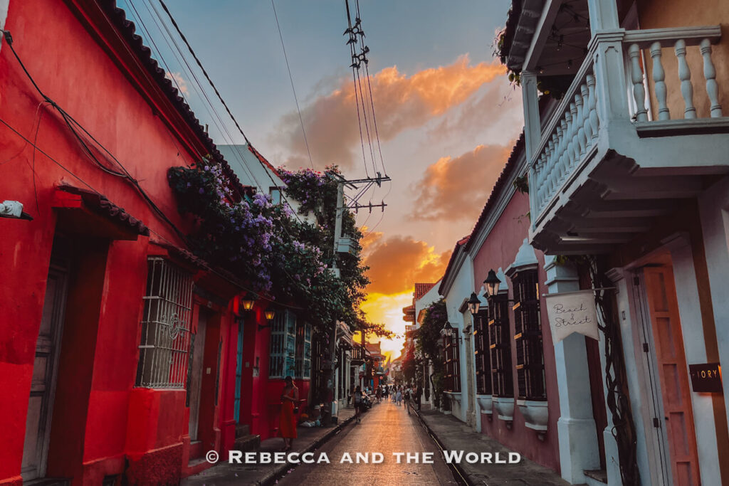 A narrow colonial street lined with colorful buildings stretches toward a glowing sunset, with pedestrians walking in the distance beneath dramatic clouds. Watching sunset in the old town is a favorite activity for visitors wondering what to do in Cartagena.