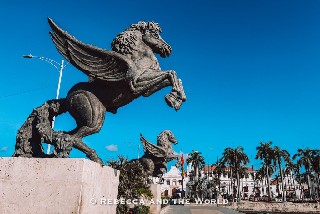 A large bronze statue of a winged horse rears up on its hind legs atop a stone pedestal, with palm trees, colonial-style buildings and a clear blue sky in the background for a Cartagena itinerary.