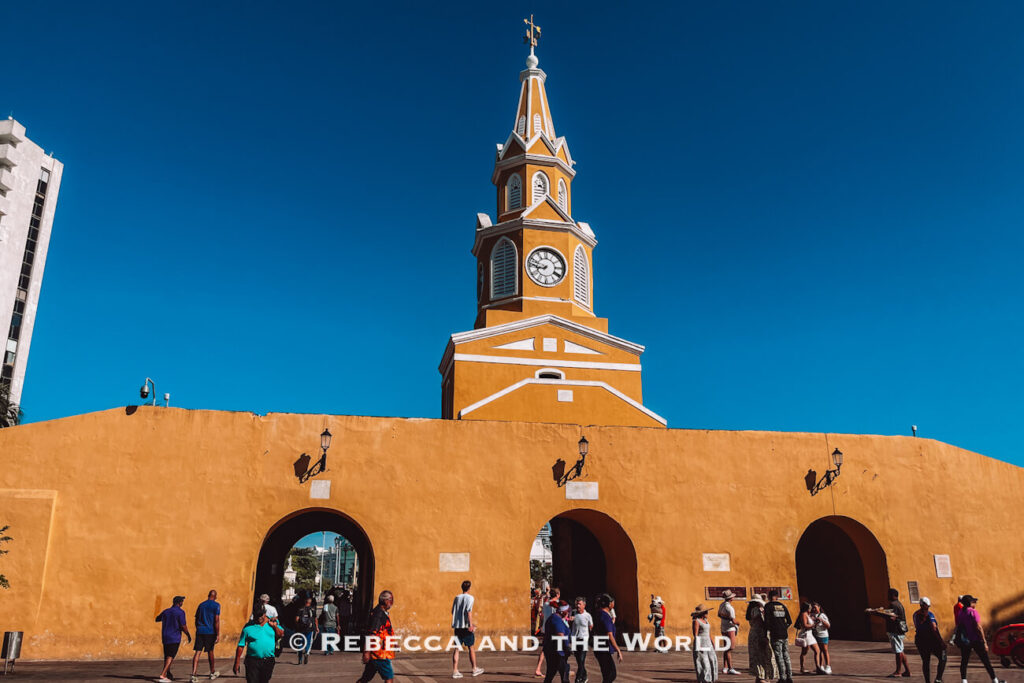 A mustard-yellow clock tower rises above a long wall with three arched openings, while people walk through the plaza during 3 days in Cartagena.