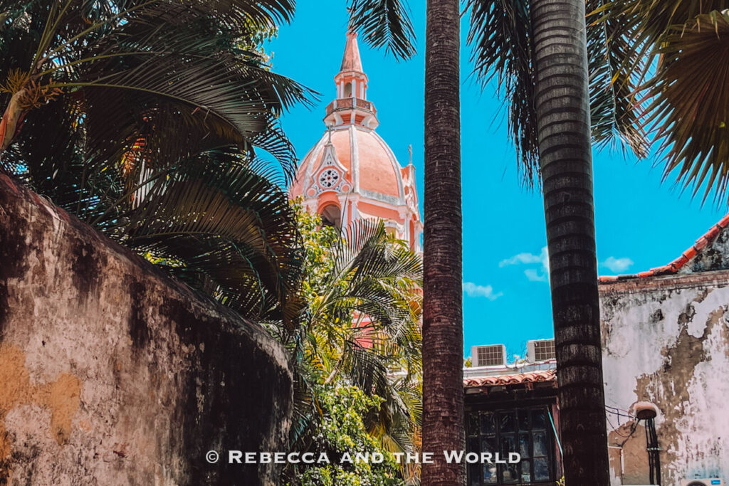 A pink-domed church tower peeks above palm trees and weathered walls in a tropical courtyard, one of the scenic things to do in Cartagena.