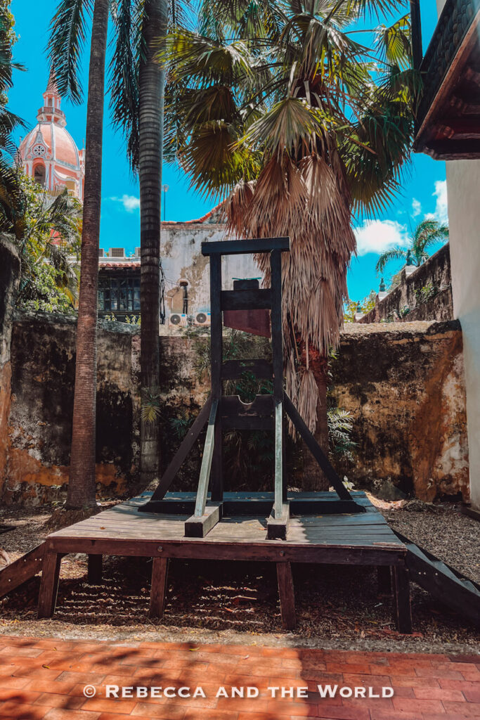 A large wooden execution device stands on a raised platform in a shaded courtyard with palm trees, old stone walls and a pink church dome, a historic stop in this Cartagena travel guide.