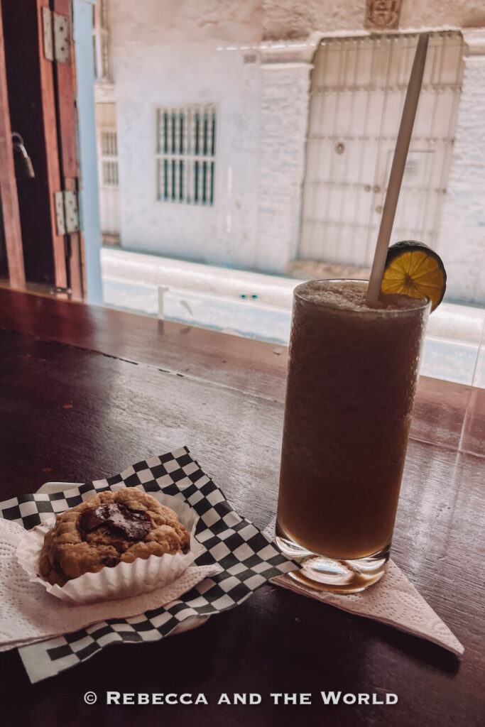 A tall iced drink with a lime slice sits beside a choc chip cookie on a wooden counter by an open window, a relaxed café stop for 3 days in Cartagena.