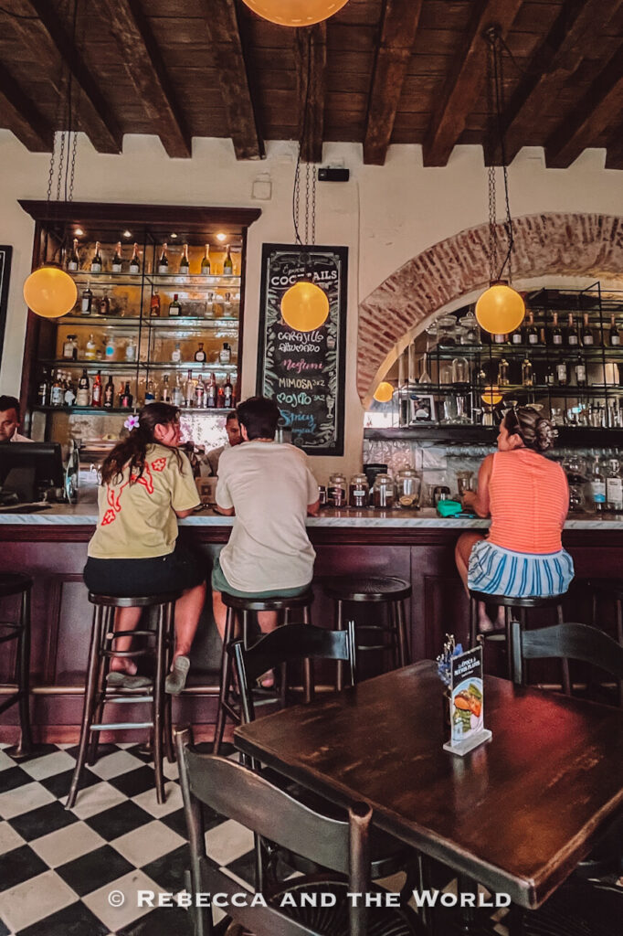 People sit at a dark wooden bar beneath globe lights and brick arches, with bottles displayed behind the counter, one of the indoor things to do in Cartagena. There are many great places to eat in Cartagena, including this bar, Epoca.