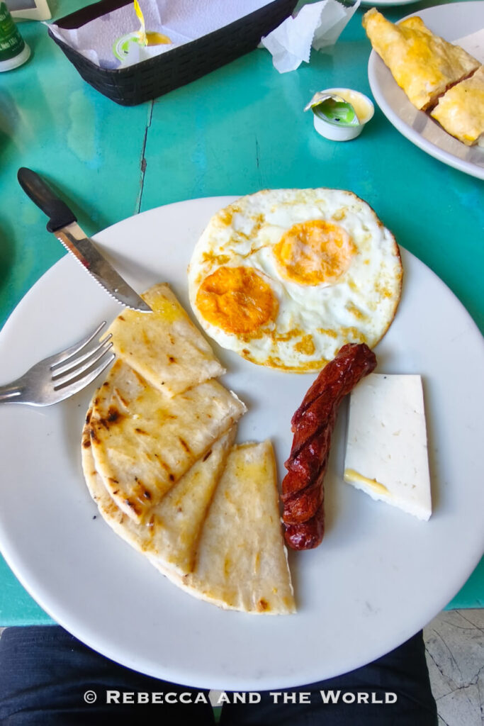 A white plate holding fried eggs, a grilled sausage, a slice of white cheese, and folded arepas sits on a turquoise table. This local breakfast is a common meal to try during 3 days in Cartagena.