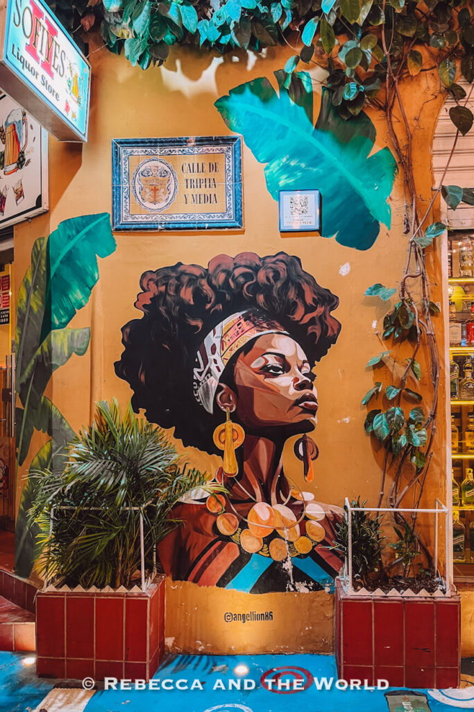 A colourful mural of a woman in large earrings and a patterned headwrap covers an ochre wall framed by tropical plants, a street-art stop for what to do in Cartagena.