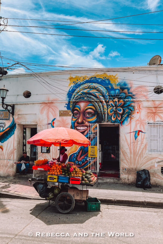 A fruit cart with a red umbrella stands in front of a large mural of a smiling woman on a white wall, a colourful street scene for a Cartagena itinerary.