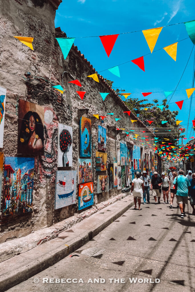 A narrow street is lined with colourful paintings and overhead red, yellow and turquoise flags while pedestrians walk by, one of the vibrant things to do in Cartagena.